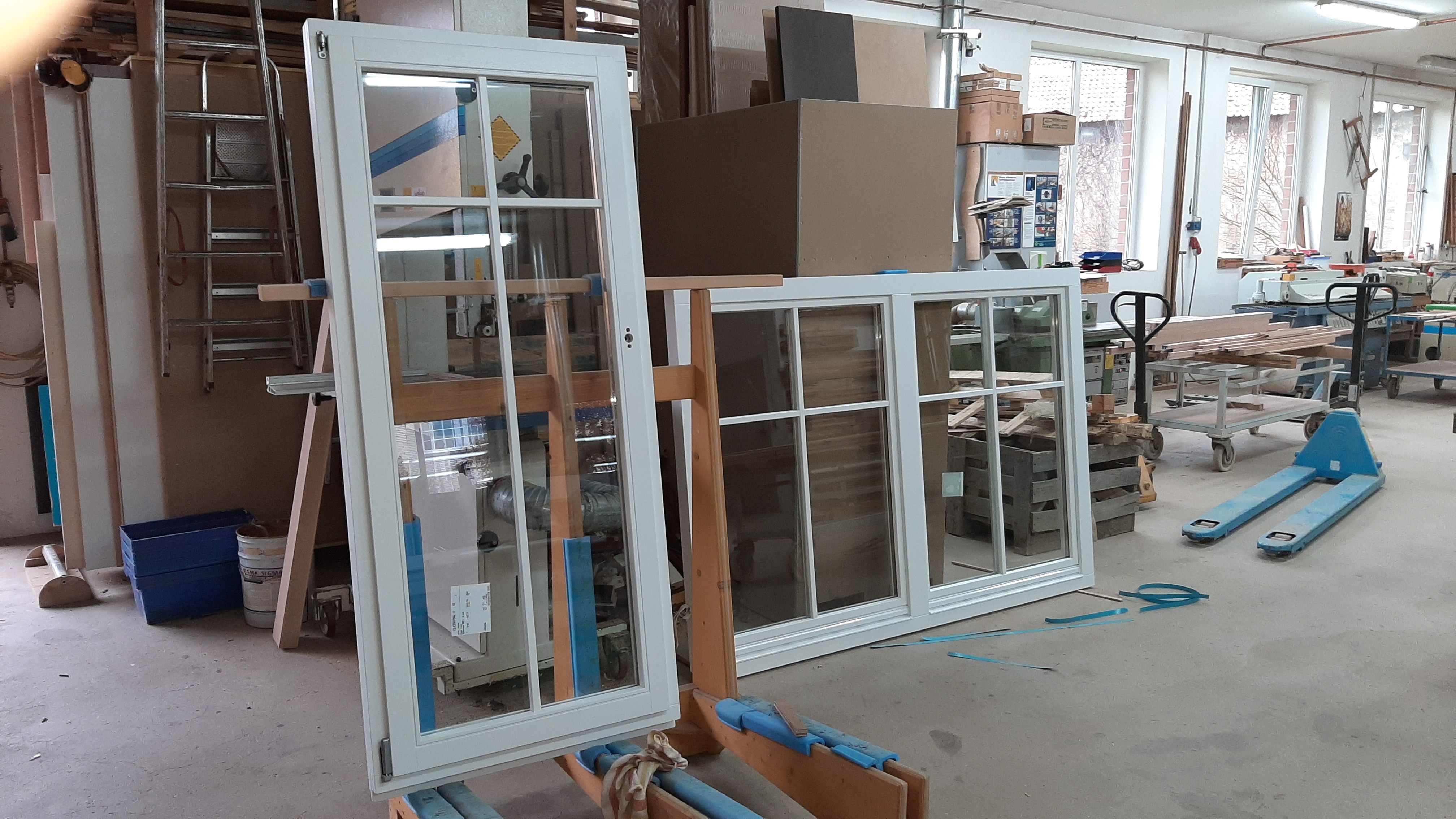 Zwei weiße Fensterrahmen in einer Werkstatt mit Kartons und Werkzeugen im Hintergrund.