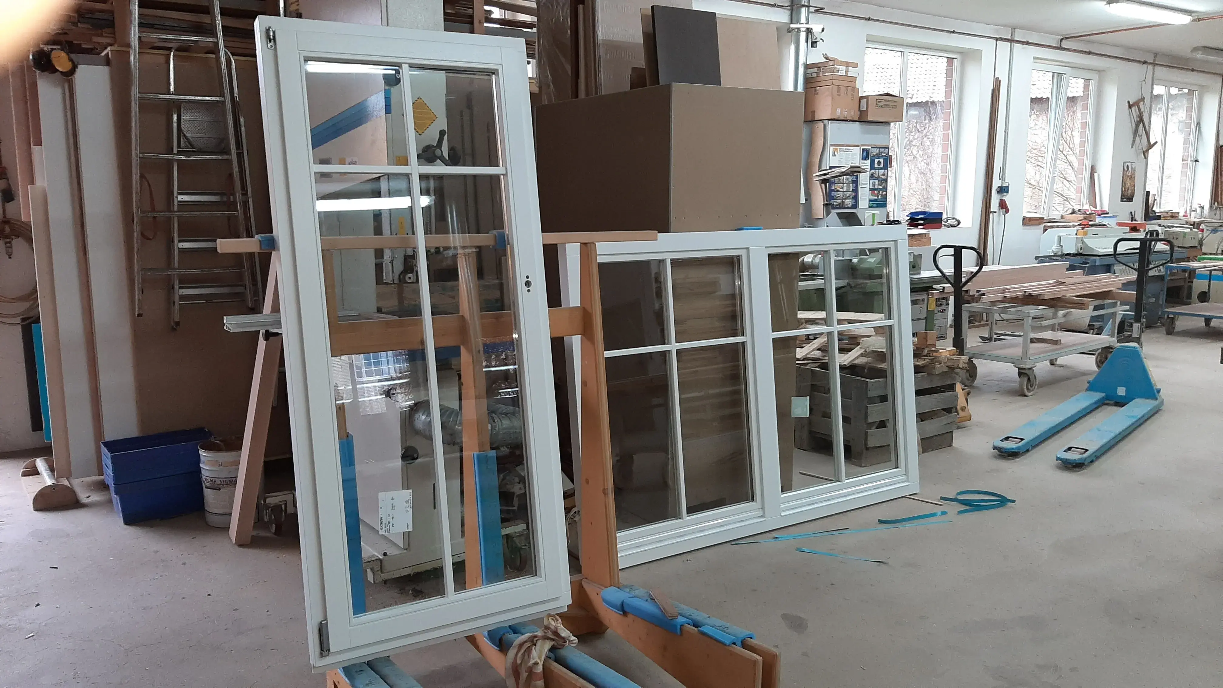 Zwei weiße Fensterrahmen in einer Werkstatt mit Kartons und Werkzeugen im Hintergrund.
